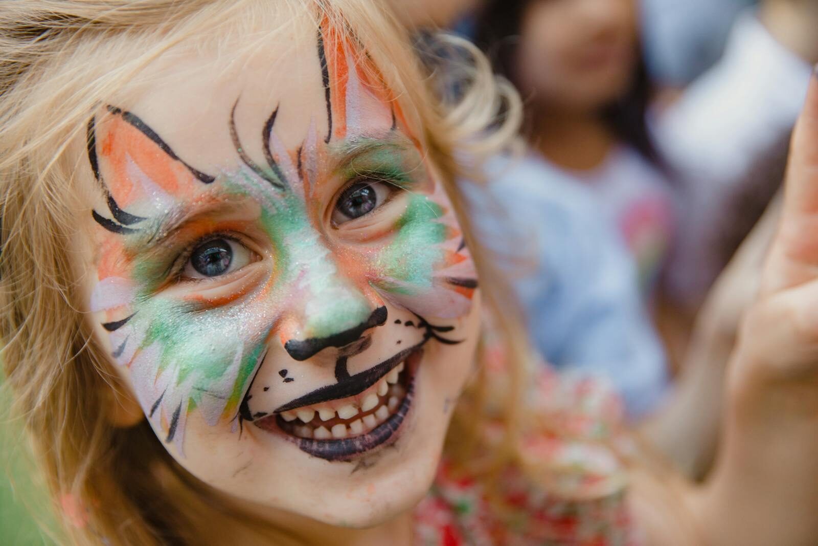 A cheerful young girl with vibrant animal face paint smiles brightly at an outdoor event.